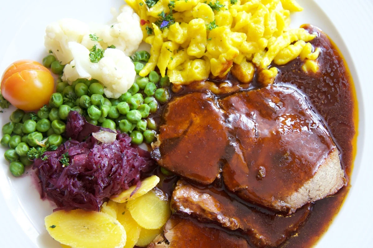 Plastry pieczonego schabu w ciemnym sosie, podane na talerzu z kolorowymi dodatkami: kluseczkami, gotowanymi ziemniakami, groszkiem, kalafiorem, czerwoną kapustą i pomidorem